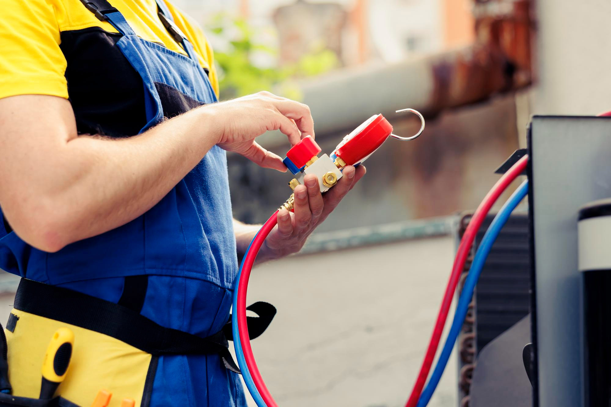 A technician in blue overalls uses a manifold gauge to service an air conditioning unit outdoors.