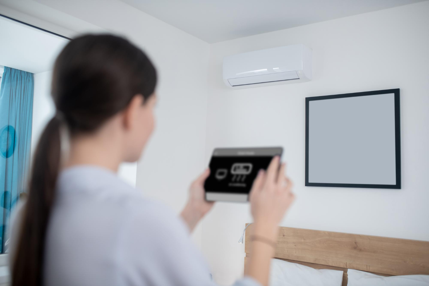 A woman uses a tablet to control a wall-mounted air conditioner in a modern bedroom.