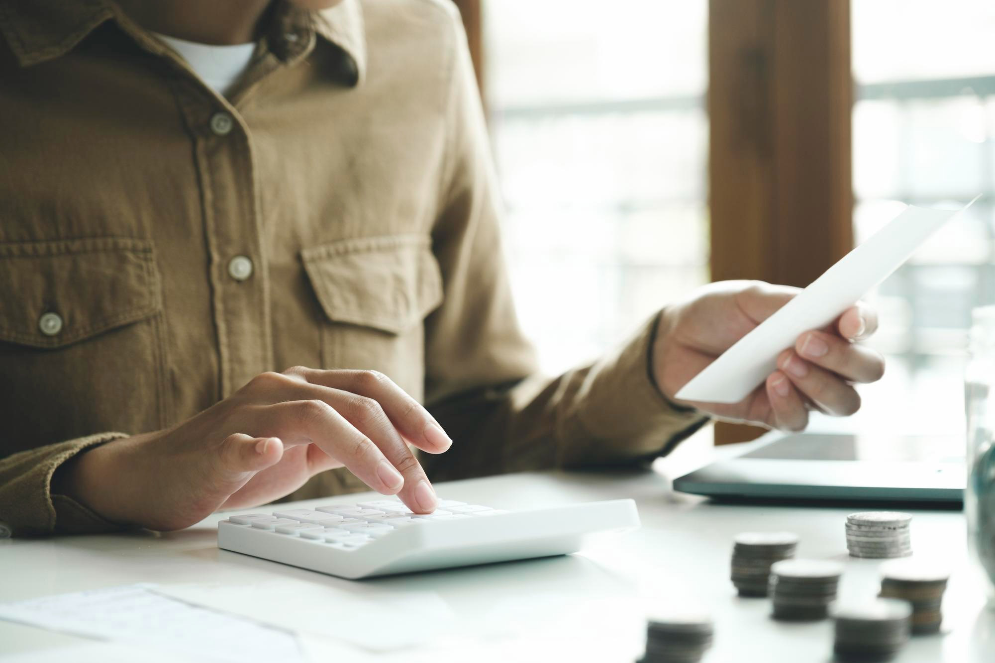 A person in a brown shirt uses a white calculator while holding a document, with stacks of coins nearby on a desk.