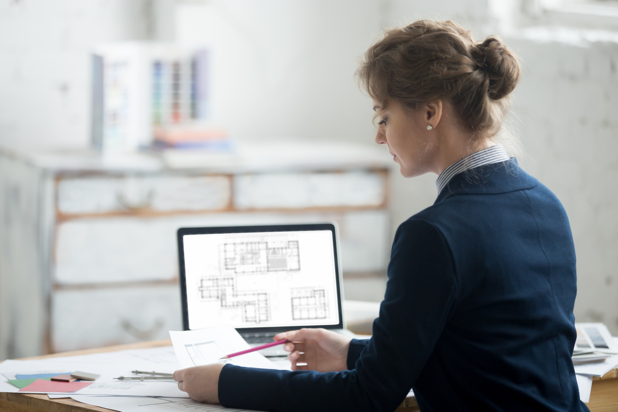 A woman in a blue blazer reviews architectural blueprints on a laptop while holding a pencil at her desk.