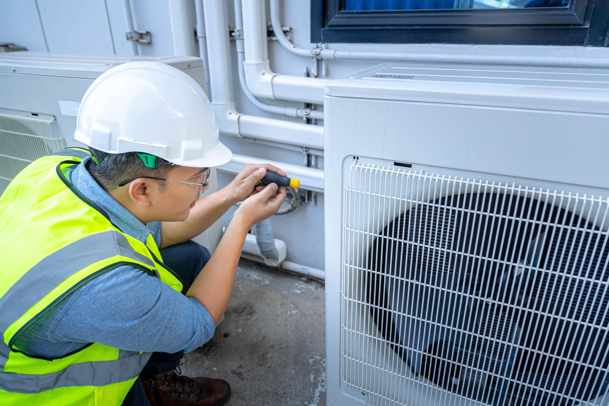 A technician in a hard hat and safety vest uses a screwdriver to adjust pipes near an outdoor air conditioning unit.