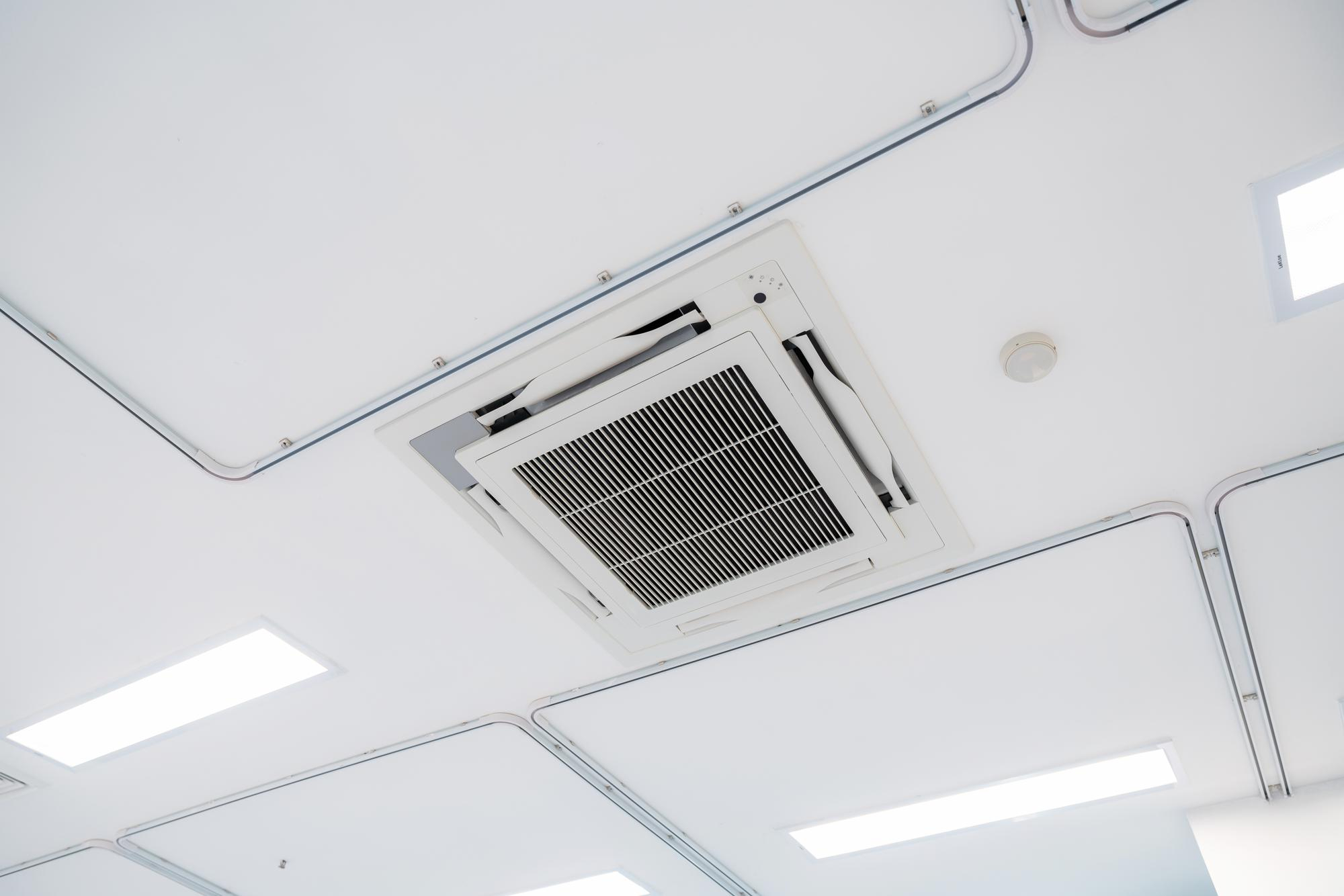 A white ceiling-mounted air conditioner unit in a modern, minimalist room with recessed lighting.