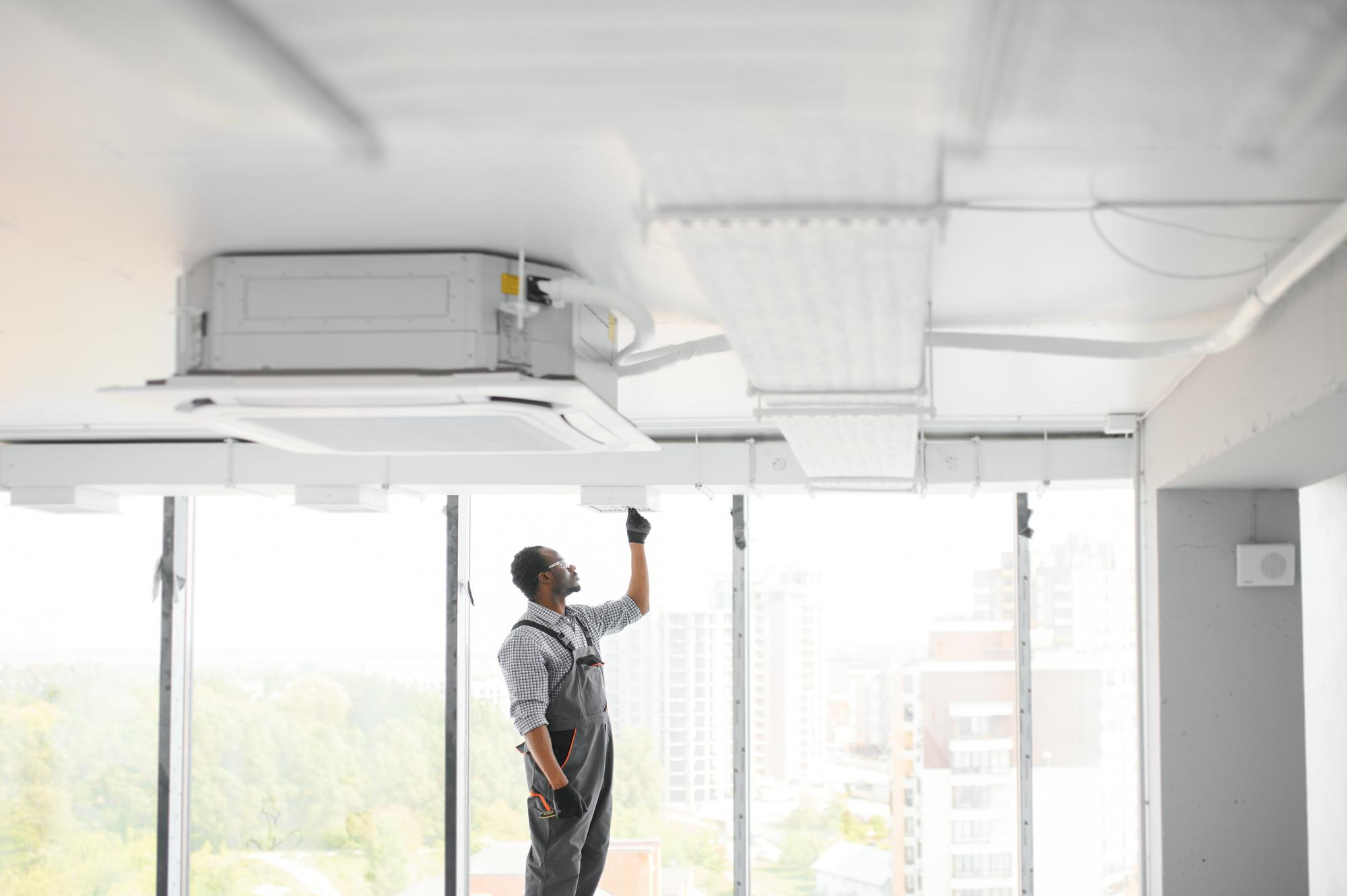 A technician in overalls installs or adjusts a ceiling-mounted HVAC unit in a modern, high-rise building with large windows.
