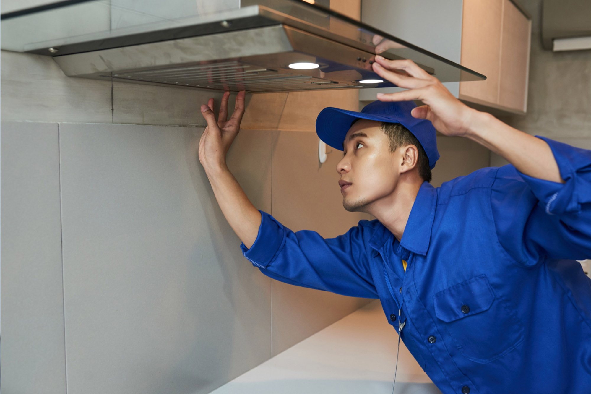 A technician in a blue uniform checks a stainless steel range hood in a modern kitchen.
